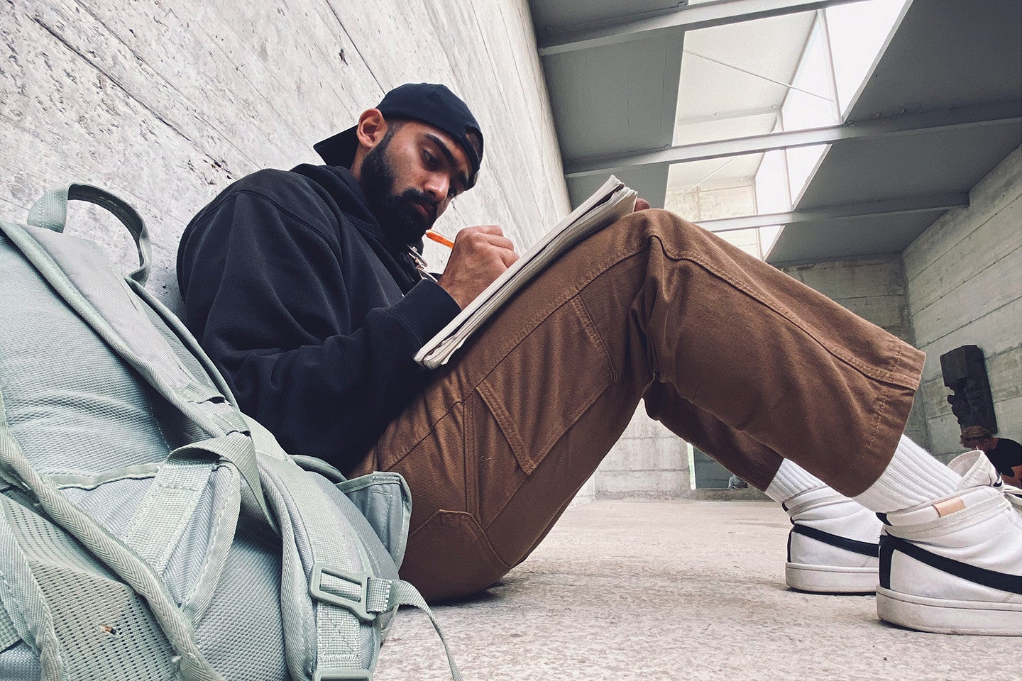 Yash sitting down in a hallway leaned against a wall writing in a notebook