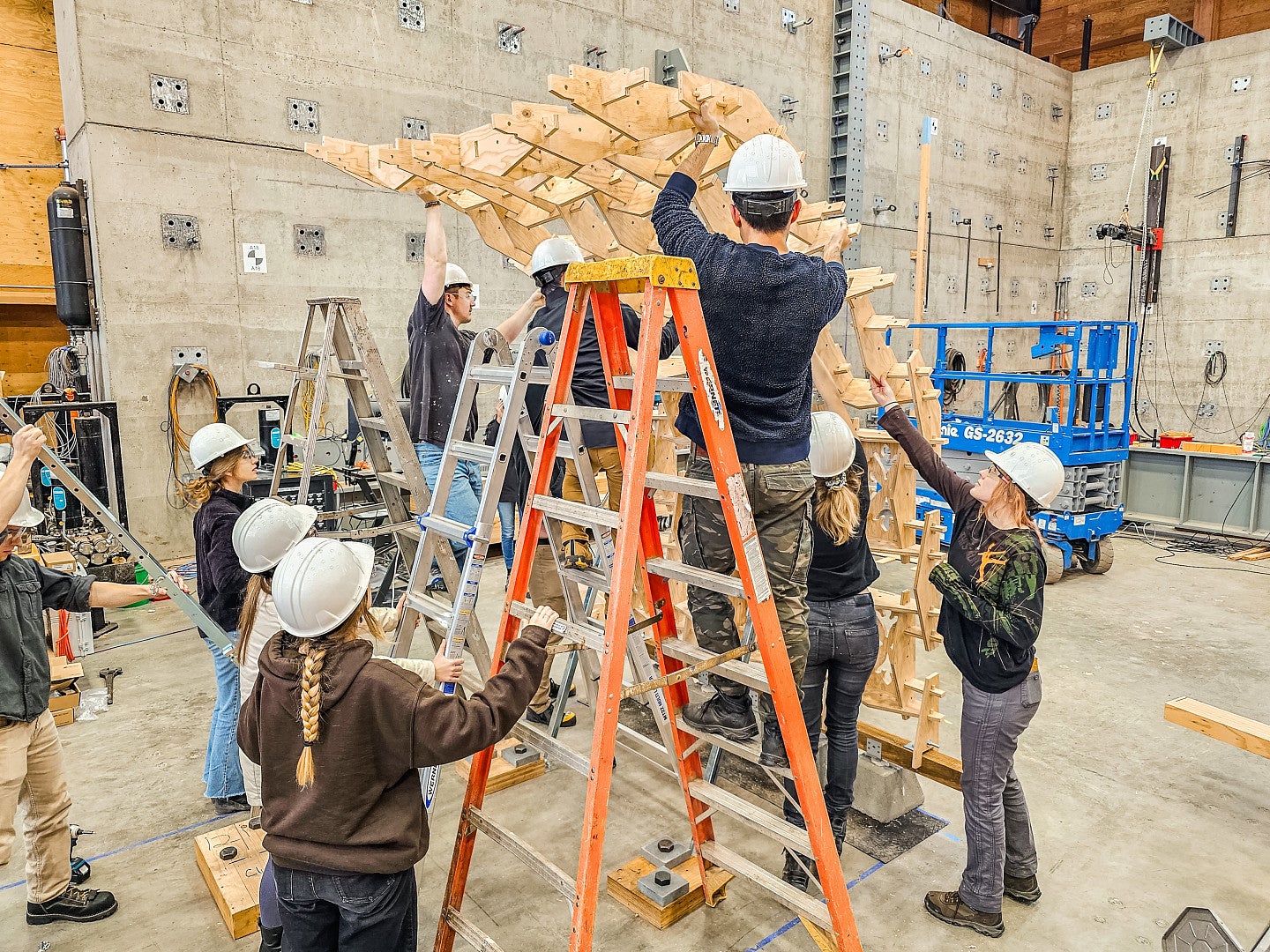 students putting first section of the arch into place. 3 students are on ladders with 6 students supporting them and the structure