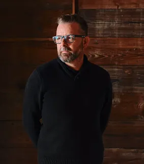 Benje Feehan looks to the left in a wooden room. Shows an older masc presenting person with glasses and a beard looking left. 