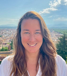 Julia Lindgren looks into the camera with a cityscape behind her. Shows a femme presenting individual with long hair and smiling. 