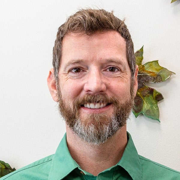 Photograph of Jeff Billington, shows a bearded male in a green button up shirt in front of a white wall with green, ceramic leaves on the back.