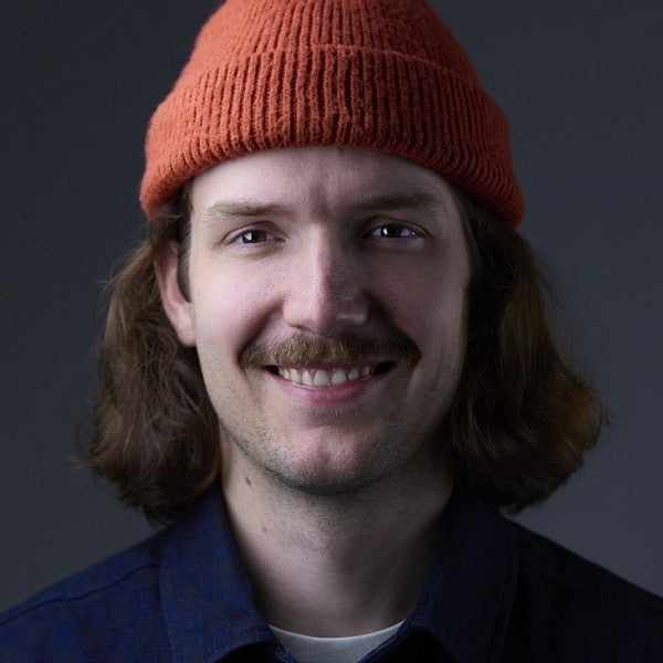 Photograph of Craig Robbins. Shows a man looking into the camera, smiling, from the shoulders up. The individual has a red-orange beanie, neck length long hair, and a smile. 