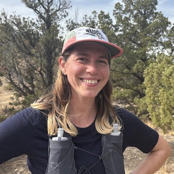 Photograph of Mica Russo. Shows a femme presenting individual in a high desert scene with shoulder length hair and smiling into the camera.
