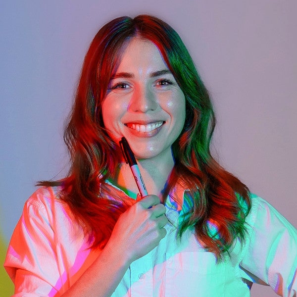 Photograph of Liz Zarro. Shows a femme presenting individual with a broad smile and posed in front of a projection of red, yellow, and cyan shadows of themselves. 