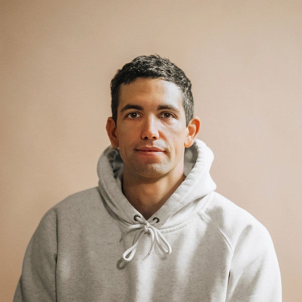 Photograph of McKenzie Sampson. Shows a young masculine person looking into the camera in front of a beige background wearing a hoodie with close cropped hair. 