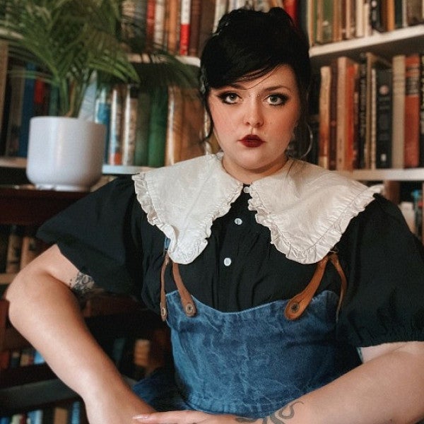 Photograph shows femme presenting person looking into the camera in a stylish, anachronistic/historic looking outfit in front of shelves of books.