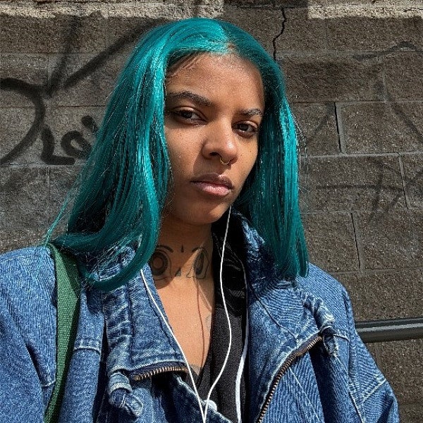 Femme presenting individual with long blue hair looks into the camera in front of a concrete wall. 