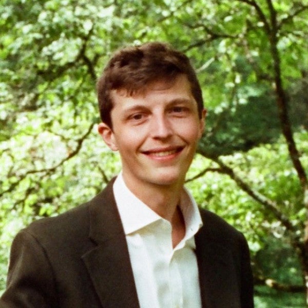 Masc-presenting individual in front of a green trees looks into the camera with a smile. 