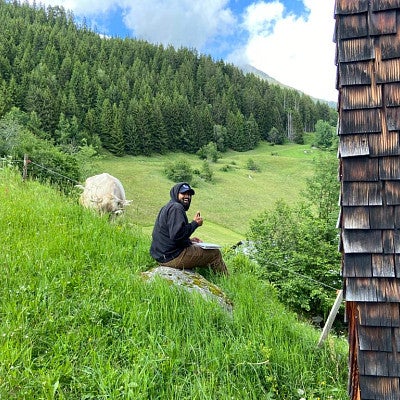 Yash giving a thumbs up, sitting in a lush field near a cow. A forest is in the distance with a bright blue sky.