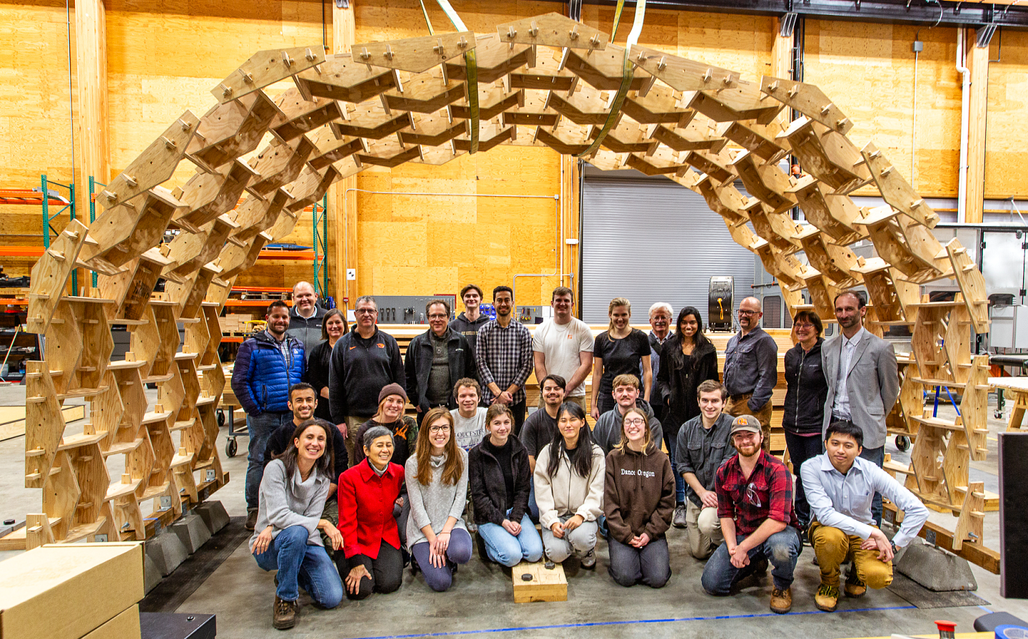 Group photo of UO and OSU students under their full-scale covered, wooden, accordion-life shelter