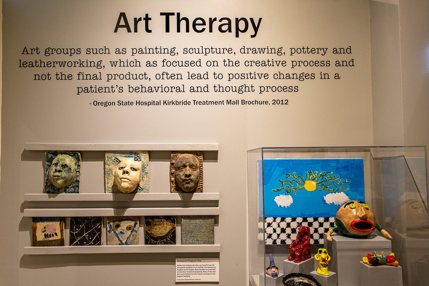 Art Therapy exhibit with multiple ceramic scupltures and paintings. There is text reading: "Art Therapy. Art groups such as painting, sculpture, drawing, pottery, and leather working, which as focused  on the creative process and not the final product, often lead to positive changes in a patients behavioral and thought process."