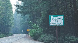 Photograph of a road that winds through tall, green trees with a "Welcome to Oregon" sign in the foreground. 