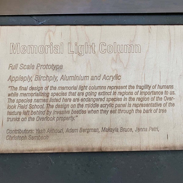 Plaque that reads, “Memorial Light Column; The final design of the memorial light columns represents the fragility of humans while memorializing species that are going extinct in regions importants to us. The species names listed here are endangered species in the region of the Overlook Field School. The design on the middle acrylic panel is representative of the texture left behind by invasive beetles when they eat through the bark of tree trunks on the Overlook Property.”